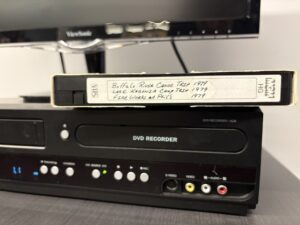 An old VHS video tape resting on a DVD player, under a monitor screen.