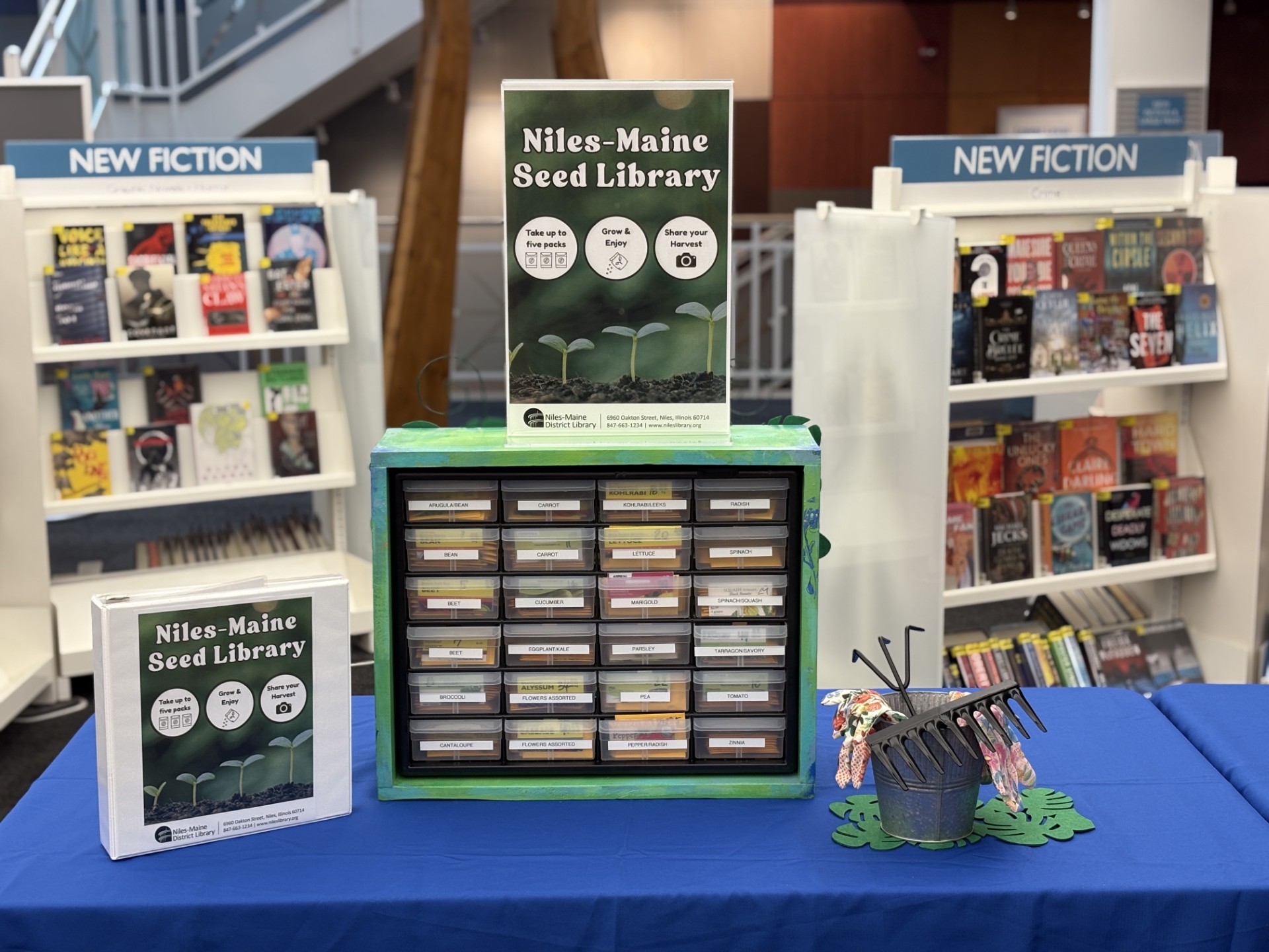 A table with a seed library and associated advertising featuring seeds for different plants for library patrons to take. In the background, library books on shelves. Image contains text that reads: Niles-Maine Seed Library on a poster and on the cover of a binder.